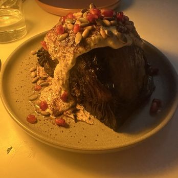 Taline - Cabbage - A plate holds a large cooked meat portion topped with sauce, seeds, and pomegranate seeds, served on a beige dish in a restaurant setting. - Mediterranean Near Me - Toronto, Ontario