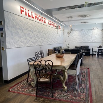 Fillmore Coffee - The photo depicts the interior of Fillmore Coffee, with a wooden dining table surrounded by ornate metal chairs placed on a patterned rug. In the background, there is a seating area with black sofas and additional chairs, along with a textured white wall and a sign reading 'FILLMORE COFFEE.' The ceiling has visible air vents and lighting fixtures, and the space includes decorative elements like plants and hanging ornaments. - Coffee & Tea Near Me - Phoenix, Arizona