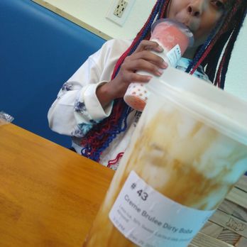 Feng Cha - A girl with multicolored braids is sitting at a table, drinking a pink beverage from a clear cup, with a blue booth and a wall outlet in the background. - Coffee & Tea Near Me - Grapevine, Texas