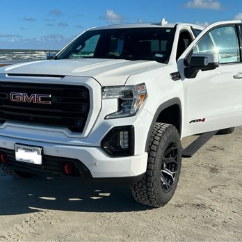 Infinity Conversions - A white GMC pickup truck is positioned on a sandy beach, with the ocean and a partly cloudy sky visible behind it. The vehicle's front and side are shown, with the driver's side door open. The truck features black wheels, a prominent grille with GMC branding, and red tow hooks on the front bumper. The scene suggests a coastal location with other vehicles visible in the distance. - Car Stereo Installation Near Me - Round Rock, Texas