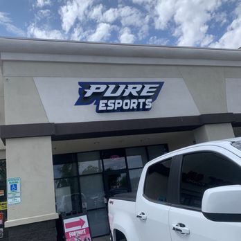 Pure Esports - Outside - The image shows the exterior of Pure Esports, a gaming center, with a large sign displaying the business name. A white pickup truck is parked in front of the entrance, which has glass doors and windows. There is a reserved parking sign for handicapped parking on the left side and a promotional sign for Fortnite with an arrow pointing toward the entrance. The sky above is partly cloudy with blue sky visible. - LAN Centers Near Me - Gilbert, Arizona