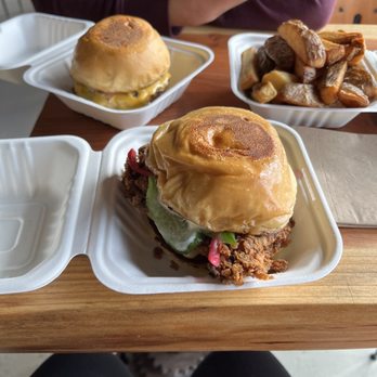 Zandos - Three takeout containers hold burgers and roasted potatoes on a wooden table, with a person in a maroon jacket visible in the background. - Food Trucks Near Me - Eugene, Oregon
