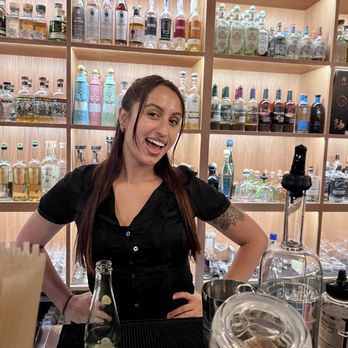 Luna Tequila - Chloe, the bartender - A woman with long brown hair, wearing a black shirt, stands behind a bar with bottles and bar tools, smiling at the camera. - Whiskey Bars Near Me - Slidell, Louisiana
