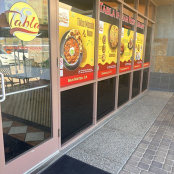 Tabla - The photo depicts the storefront of Tabla Indian Restaurant, featuring large glass windows with promotional posters advertising dishes such as Tikka Masala and naan. The posters include the restaurant's locations in San Mateo, CA, Danville, CA, and Pleasanton, CA. The entrance has a glass door with a metal handle, and the sidewalk outside is paved with bricks and concrete. The interior of the restaurant is partially visible through the glass. - Indian Near Me - Phoenix, Arizona