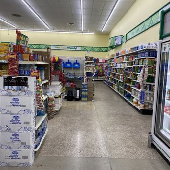 Dollar Tree - The image shows an aisle inside a Dollar Tree store with shelves on both sides filled with various products, including snacks, household supplies, and holiday decorations. The ceiling has fluorescent lighting, and the floor is made of speckled tiles. The aisle appears organized, with some items stacked on pallets and boxes at the end of the aisle. The store signage indicates weekly deals and new items. - Discount Store Near Me - Placentia, California