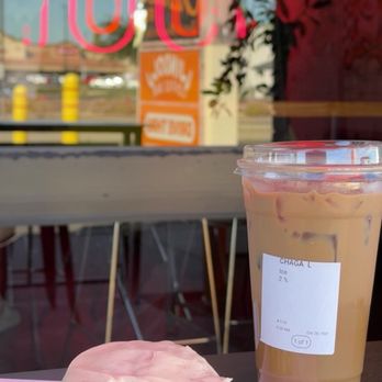 Window Coffee Bar - Chagaccino and scone - A cookie with pink icing is on a pink paper sheet on a table, alongside a large iced coffee in a clear plastic cup with a lid. The coffee has a label indicating it is a Chaga L with 2% ice. The background shows a window with reflections and a view of the outside, including a sign and some yellow posts. - Coffee & Tea Near Me - Phoenix, Arizona