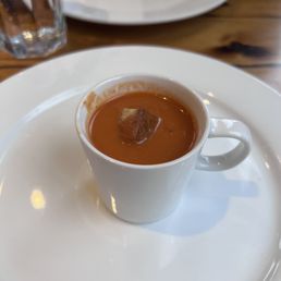Tomato soup - A white ceramic cup filled with tomato soup, garnished with a small piece of bread or crouton, placed on a white plate on a wooden table.