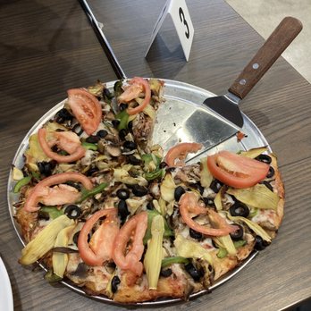 Mountain Mike's Pizza - Mt. Veggiemore - A vegetable pizza with toppings including tomato slices, black olives, green peppers, artichoke hearts, and mushrooms on a metal tray at a restaurant. - Chicken Wings Near Me - Greenfield, Wisconsin