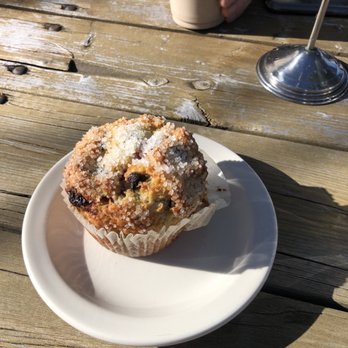 Velo Cafe - Raspberry chocolate chip muffin. Gluten free but you couldn't tell! - A muffin topped with sugar crystals sits on a white plate on a weathered wooden table, with a beverage and a metal stand nearby. - Juice Bars & Smoothies Near Me - Cable, Wisconsin