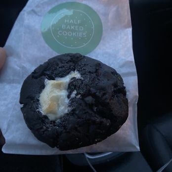 Half Baked Cafe - Hot Chocolate - A dark chocolate cookie with a slightly cracked surface and a piece of butter on top is held in hand over a paper bag from Half Baked Cookies. The background shows the interior of a vehicle, including part of a dashboard and a cup holder. The cookie appears freshly baked and rich in chocolate. - Ice Cream & Frozen Yogurt Near Me - Lockport, New York