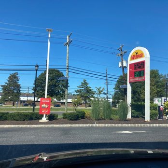 Wawa - a gas station with a large sign - The image features a Wawa gas station sign displaying fuel prices, with a bright blue sky and some trees in the background. The sign is situated on a landscaped area with bushes and grass, and there are utility poles and power lines visible above. The scene appears to be taken from inside a vehicle, with part of the dashboard or windshield at the bottom of the image. - Gas Stations Near Me - Hamilton, New Jersey