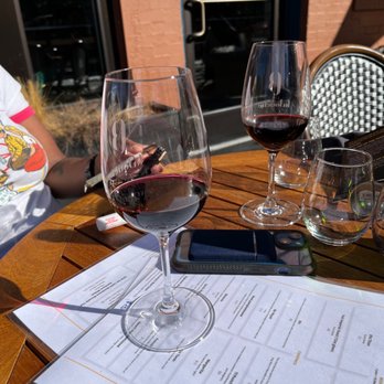 La Bouche - Two glasses of red wine are placed on a wooden table outside a restaurant, with a menu, a smartphone, and a glass of water nearby. - Wine Bars Near Me - Denver, Colorado