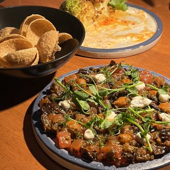 Ossobuco Miami- Steak house - tartare with eggplant - A meal with three dishes and a bowl of chips on a wooden table, including a vegetable dish, a creamy dish, and a roasted vegetable plate. - Steakhouses Near Me - Miami, Florida