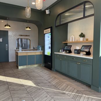 Wonder Cresskill - The image shows the interior of a modern cafe or restaurant with a green wall and wooden flooring. There is a counter with a mirror and a vending machine nearby. Two self-order kiosks are positioned on a green cabinet with a wooden shelf above, decorated with a potted plant and small decorative items. Pendant lights hang from the ceiling, and an exit sign is visible. The space appears clean and well-lit. - Food Delivery Services Near Me - Cresskill, New Jersey