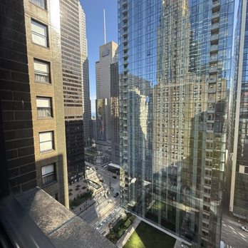 Pendry Chicago - Photograph captures a city scene from a high vantage point, showing multiple tall skyscrapers with glass facades reflecting neighboring buildings and the blue sky. The street below has some vehicles and pedestrians, with a small green space visible at the bottom. The reflections create a layered visual effect of the urban environment. - Hotels Near Me - Chicago, Illinois