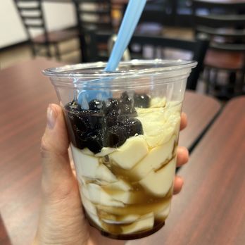 Scooptopia - Taho - A person holds a clear plastic cup filled with halo halo, a Filipino dessert, featuring beans, ice cream, and syrup. The background shows a cafe interior with wooden tables and chairs, some of which are empty. The person’s hand is visible, and a light blue spoon is inserted into the dessert. - Ice Cream & Frozen Yogurt Near Me - Phoenix, Arizona