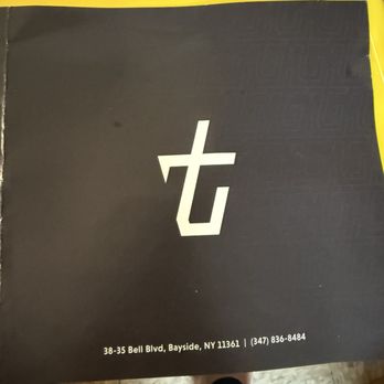 Tbaar - Bayside - The image shows a black menu or booklet with a white stylized logo on the front cover. The logo appears to be a lowercase 't' and 'b' combined. The bottom of the cover displays the address '38-35 Bell Blvd, Bayside, NY 11361' and a contact phone number '(347) 836-8484'. The background includes a yellow surface and a person's legs and feet visible at the bottom of the image, indicating the photo was taken from a standing position. - Bubble Tea Near Me - Bayside, New York