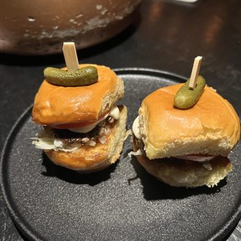BarChef - Two mini sliders with pickles on top are placed on a black plate, with visible layers of meat, vegetables, and bread. - Lounges Near Me - Toronto, Ontario