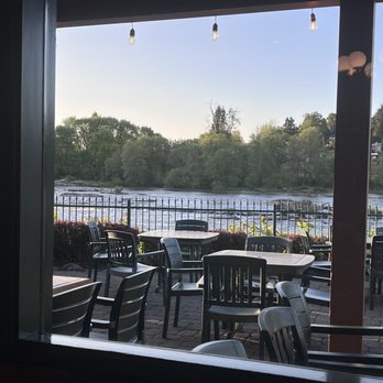 Roaring Rapids Pizza Co - Beautiful flowing river outside - View through a window showing an outdoor patio with multiple tables and chairs, situated near a river. The patio is bordered by a fence and surrounded by greenery, including trees and bushes. Overhead string lights are visible, and the sky is clear, indicating a bright day. The setting suggests a riverside restaurant or cafe with a scenic outdoor seating area. - Pizza Near Me - Eugene, Oregon