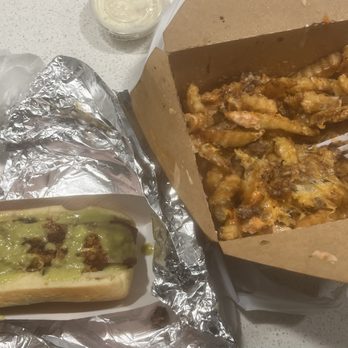 DittyDog - The image shows a hot dog in a paper tray, topped with melted cheese and meat, with some sauce visible. Next to it is a large portion of fries served in a cardboard container, with some sauce and toppings on the fries. The food appears to be takeout, placed on a light-colored surface. - Hot Dogs Near Me - Austin, Texas