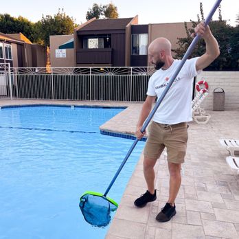 Garibaldi Pools - A bald man in a white shirt and khaki shorts cleans a blue swimming pool with a long-handled skimmer on a tiled deck. - Pool Cleaners Near Me