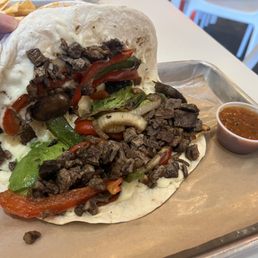 Hybrid premium Taco - A sizable taco with a soft flour tortilla contains grilled beef, red and green peppers, onions, and lettuce, accompanied by a small cup of salsa on a metal tray.