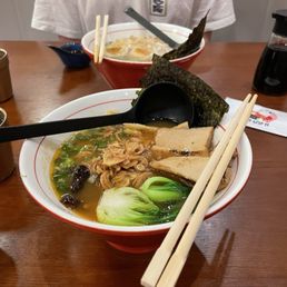 Vegan ramen - Two bowls of ramen are placed on a wooden table, each containing broth, noodles, seaweed, slices of pork, and vegetables. Chopsticks rest on the bowls, and spoons are included for eating. In the background, a person wearing a white shirt is partially visible, and condiments are on the table.