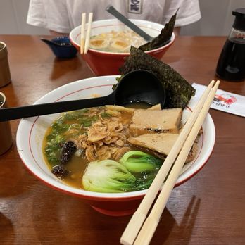 Umami - Vegan ramen - Two bowls of ramen are placed on a wooden table, each containing broth, noodles, seaweed, slices of pork, and vegetables. Chopsticks rest on the bowls, and spoons are included for eating. In the background, a person wearing a white shirt is partially visible, and condiments are on the table. - Ramen Near Me - Annapolis, Maryland