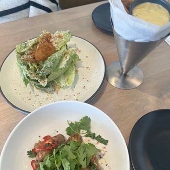 Lynette - The photo features a meal with three main items: a Caesar salad with lettuce, croutons, and grated cheese on a white plate; fried chicken wings topped with cilantro and sliced red chili peppers on a white dish; and a serving of French fries in a metal cone lined with parchment paper, accompanied by a small container of dipping sauce, likely cheese or aioli. The setting appears to be a casual dining environment with a wooden table. - Beer Bar Near Me - Minneapolis, Minnesota