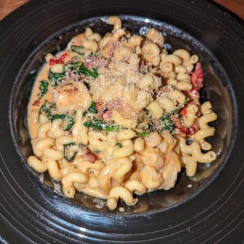 Table 32 - Seafood Mac and Cheese - A bowl of creamy macaroni and cheese with spinach, bacon, and breadcrumbs, served in a black dish on a wooden table. - New American Near Me - Sioux City, Iowa