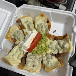 a tray of nachos with cheese and sauce - A foam takeout container contains nachos with melted cheese, guacamole, sour cream, a tomato slice, and shredded beef, served as a Mexican snack or meal.