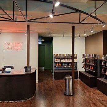 Hotel Chocolat - The image shows the interior of a modern chocolate shop with shelves of chocolate products, a curved counter with registers, and a neon sign on the wall. - Chocolatiers & Shops Near Me - Chicago, Illinois