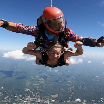 Jumptown Skydiving - A woman and an instructor are skydiving in tandem, with the instructor wearing a red helmet and the woman smiling, high above a landscape of fields and forests. - Skydiving Near Me - Orange, Massachusetts