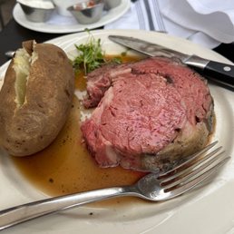 Sir Lancelot Cut - A white plate holds a cooked steak with visible juices, accompanied by a baked potato with a split open top, and a small bunch of microgreens. A knife rests on the plate, and a fork is placed nearby. In the background, there are small containers of sauces and condiments, and a white napkin. The setting appears to be a restaurant with a dark table surface and additional dishes visible.