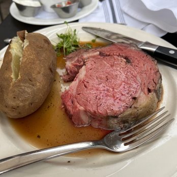 Lancelot Restaurant - Sir Lancelot Cut - A white plate holds a cooked steak with visible juices, accompanied by a baked potato with a split open top, and a small bunch of microgreens. A knife rests on the plate, and a fork is placed nearby. In the background, there are small containers of sauces and condiments, and a white napkin. The setting appears to be a restaurant with a dark table surface and additional dishes visible. - Steakhouses Near Me - Vail, Colorado