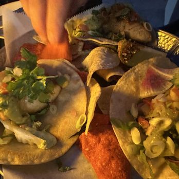 Pesca Vilano - Fish tacos - A close-up image shows a plate of fish tacos with various toppings including green onions, cilantro, and possibly diced vegetables. A hand is seen adding garnishes or toppings to the tacos. The scene appears to be in a dimly lit setting, likely at a restaurant or outdoor dining area, with the focus on the food presentation. - Seafood Near Me - St. Augustine, Florida