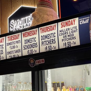 Wings Etc - Weekly drink specials - A menu board lists weekly drink specials at a bar, including prices for domestic bottles, pints, tankers, schooners, pitchers, and light or MG pitchers. The board is illuminated and positioned above a glass-front refrigerator containing bottled beverages. A Santa hat is placed on top of the board, and a neon sign with the word 'SAMUEL' is visible in the background. - Sports Bars Near Me - Crystal Lake, Illinois