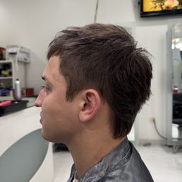 A person with a short, layered hairstyle is seated in a salon chair, wearing a black salon cape, in a hair salon setting.
