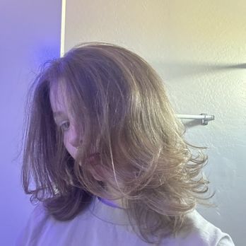 Precision Cut - A person with shoulder-length, wavy hair that partially covers their face is shown in an indoor environment. They are wearing a light-colored shirt, and the background includes a textured wall and a towel rack. The lighting creates a soft, neutral tone in the image. - Hair Salons Near Me - Gilbert, Arizona