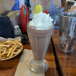 Chocolate Coconut Shake - A chocolate coconut milkshake with whipped cream and a cherry on top is served in a tall glass on a wooden table, with fries and condiments nearby.