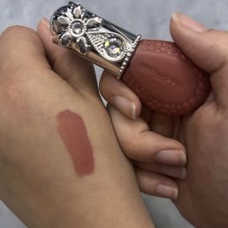 Jill Stuart Bloom Droff No. 3 - A hand is gripping a lipstick with a decorative, jeweled cap featuring floral and gemstone details. A swatch of lipstick is applied on the person's arm, showing the color and texture of the product. The background is neutral and out of focus.