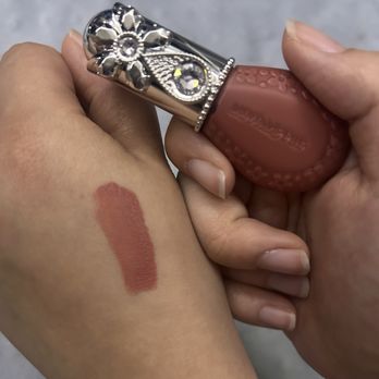 Maison Kose - Jill Stuart Bloom Droff No. 3 - A hand is gripping a lipstick with a decorative, jeweled cap featuring floral and gemstone details. A swatch of lipstick is applied on the person's arm, showing the color and texture of the product. The background is neutral and out of focus. - Cosmetics & Beauty Supply Near Me - Arcadia, California