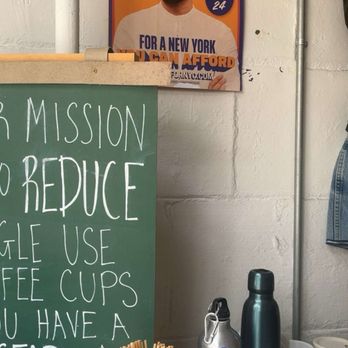IXV Coffee - A campaign poster for Zohran Mamdani, a Democratic candidate, is taped to a white wall in a coffee shop. The poster features a portrait of Zohran Mamdani with text promoting his candidacy for a New York election, including a vote date of June 24. Below the poster, there is a chalkboard menu with handwritten instructions about reducing single-use coffee cups, and various coffee mugs and bottles are placed on the counter. - Coffee & Tea Near Me - Brooklyn, New York