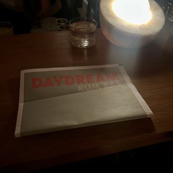 Daydream - The image shows a wooden table with a printed menu from Daydream Rum Bar. The menu is partially visible, with bold red and beige text. A salt lamp emitting a warm glow is positioned on the right side of the table, and there are glasses of water in the background. The setting appears to be dimly lit, likely in a bar or restaurant environment. - Cocktail Bars Near Me - Albuquerque, New Mexico