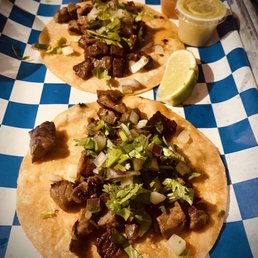 Steak Tacos - Two beef tacos with grilled beef chunks, chopped onions, and cilantro are placed on a blue and white checkered paper tray. Each taco is served on a soft corn tortilla. Accompanying the tacos are lime wedges, a small container of green sauce, and a small container of reddish-brown salsa. The presentation suggests a casual dining setting, typical of Mexican street food or taqueria style.