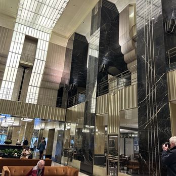Inside Chicago Walking Tours - The image shows a spacious lobby with black marble columns and high ceilings. Illuminated panels are present on the ceiling, and there are seating areas with sofas. Several people are seen walking or sitting, and a person is taking a photo. The design includes metallic accents and glass doors, indicating a modern, upscale interior. - Architectural Tours Near Me - Chicago, Illinois