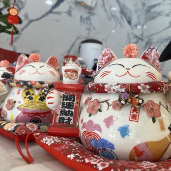 Tsaocaa - St Johns - Two ceramic lucky cat figurines with floral patterns, pink ears, and red accents are shown. They have closed eyes and smiling faces, with decorative collars and small ornaments. The figurines are placed on a surface with a marble-patterned background and a small plant visible in the corner. The display appears to be part of a decorative or festive setting, possibly related to Japanese or Asian cultural themes. - Desserts Near Me - Saint Johns, Florida