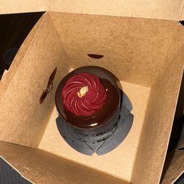 A round chocolate cake with glossy chocolate glaze and red frosting decoration, topped with gold leaf, inside a brown cardboard takeout box.
