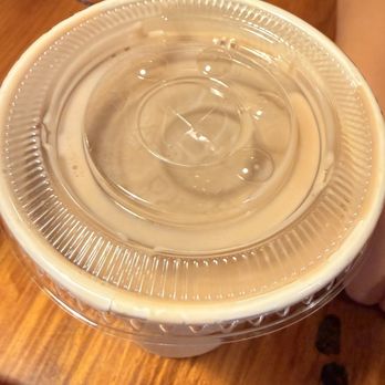 Rainbow Oaks Restaurant - Chocolate milk - A transparent plastic cup filled with chocolate milk is positioned on a wooden table. The cup has a clear plastic lid, and the milk appears smooth and creamy. In the background, there is a white napkin and some indistinct objects, suggesting a casual dining setting. - American Near Me - Fallbrook, California