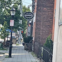 Sign outside - The image shows a city sidewalk adjacent to a brick building with a hanging sign for Gypsy's. The sidewalk has street lamps, trees, and outdoor seating with chairs and tables. Utility poles and wires are visible in the background, along with other buildings and street signs. The scene appears to be in a downtown or commercial area with pedestrian amenities.
